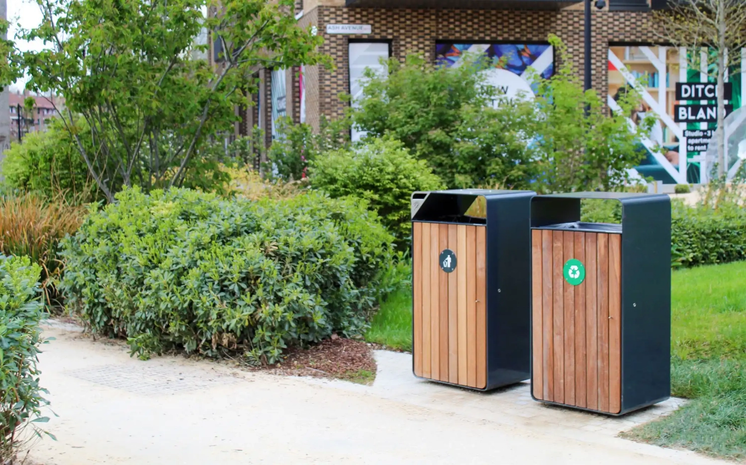 Zwei moderne Abfallbehälter mit Holzverkleidung und Recycling-Symbolen stehen in einer begrünten Umgebung mit Sträuchern und kleinen Bäumen.