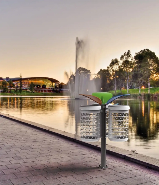 Modernes Abfallbehälter-Design am Ufer eines ruhigen Sees, im Hintergrund ein Springbrunnen und eine große Gebäude-Silhouette bei Sonnenuntergang.