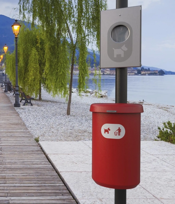 Ein roter Mülleimer für Hundekot, montiert an einem Pfosten an einer Uferpromenade, mit einem Hinweis auf Hundebesitzer. Im Hintergrund sind Bäume und das Wasser sichtbar.