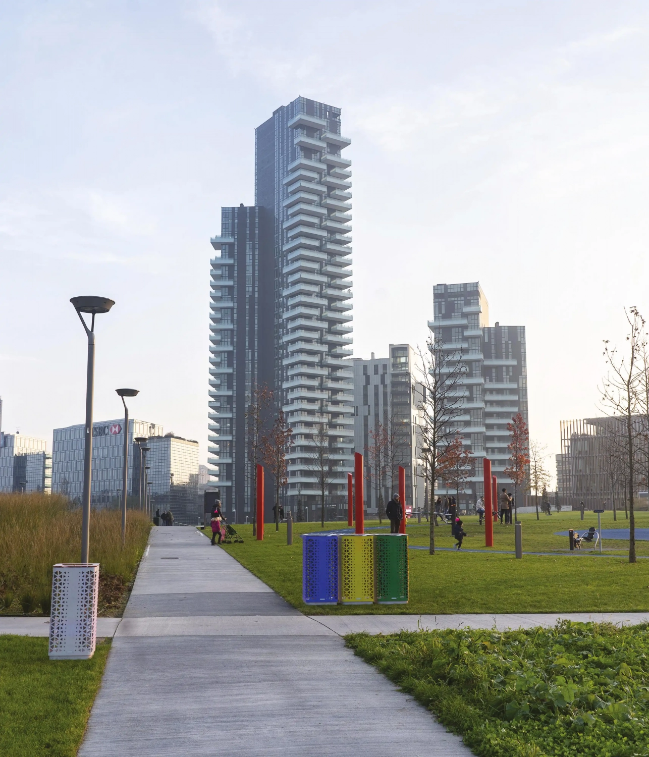 Moderne Architektur in einem urbanen Park mit Hochhäusern im Hintergrund. Zu sehen sind grüne Rasenflächen, Gehwege und farbige Mülltonnen. Einige Menschen bewegen sich im Park.