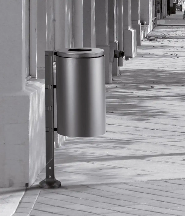 Ein moderner, silberner Mülleimer, der an einer Wand befestigt ist, steht auf einem gepflasterten Gehweg. Die Umgebung ist in Schwarz-Weiß gehalten.