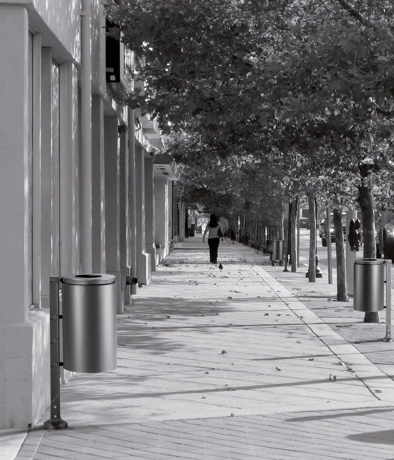 Eine schwarz-weiße Straßenansicht mit einer gepflasterten Gehweg, der von Bäumen gesäumt ist. Im Hintergrund joggt eine Person. Mülltonnen stehen entlang des Gehwegs, und einige Blätter liegen auf dem Boden.