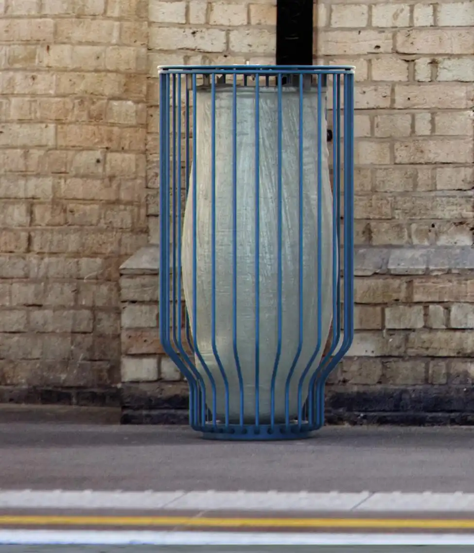 Eine große, gekörnte Vase, die von einem blauen Metallgitter umgeben ist, steht vor einer Backsteinmauer.