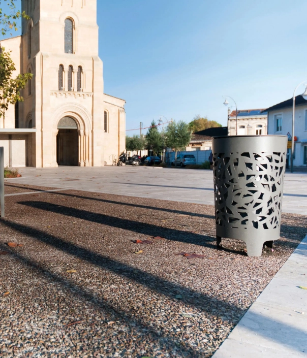 Ein moderner, metallener Abfallbehälter mit einem dekorativen Muster steht auf einem gepflasterten Platz vor einer Kirche. Im Hintergrund sind Bäume und parkende Autos sichtbar, während der Himmel klar und blau ist.