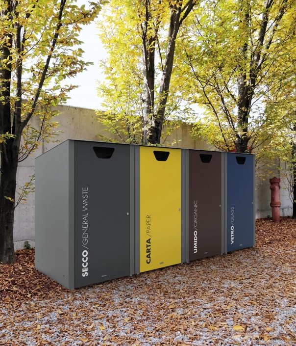 Drei Müllcontainer in verschiedenen Farben stehen vor einer Wand aus Beton, umgeben von Bäumen mit gelben Blättern im Herbst. Der erste Container ist für Restmüll, der zweite für Papier, der dritte für organische Abfälle und der letzte für Glas.