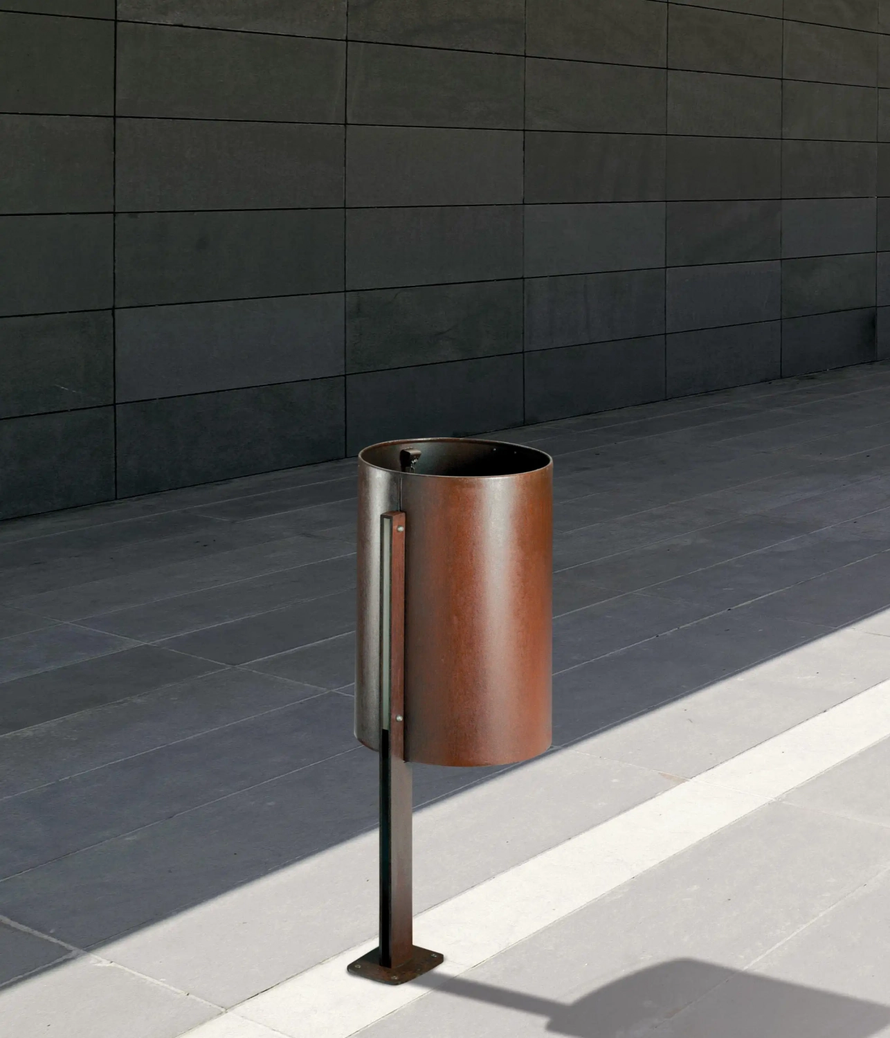 corte-abfallbehaelter-03 Ein moderner, runder Mülleimer aus Metall steht auf einem grauen Platz vor einer schlichten, dunklen Wand.