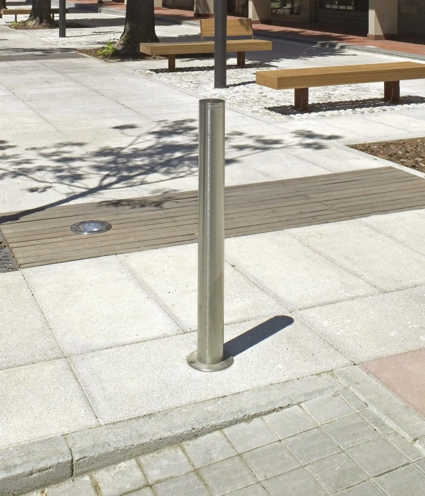 Edelstahlpoller auf einem Fußweg, umgeben von Pflastersteinen und Holzbank.