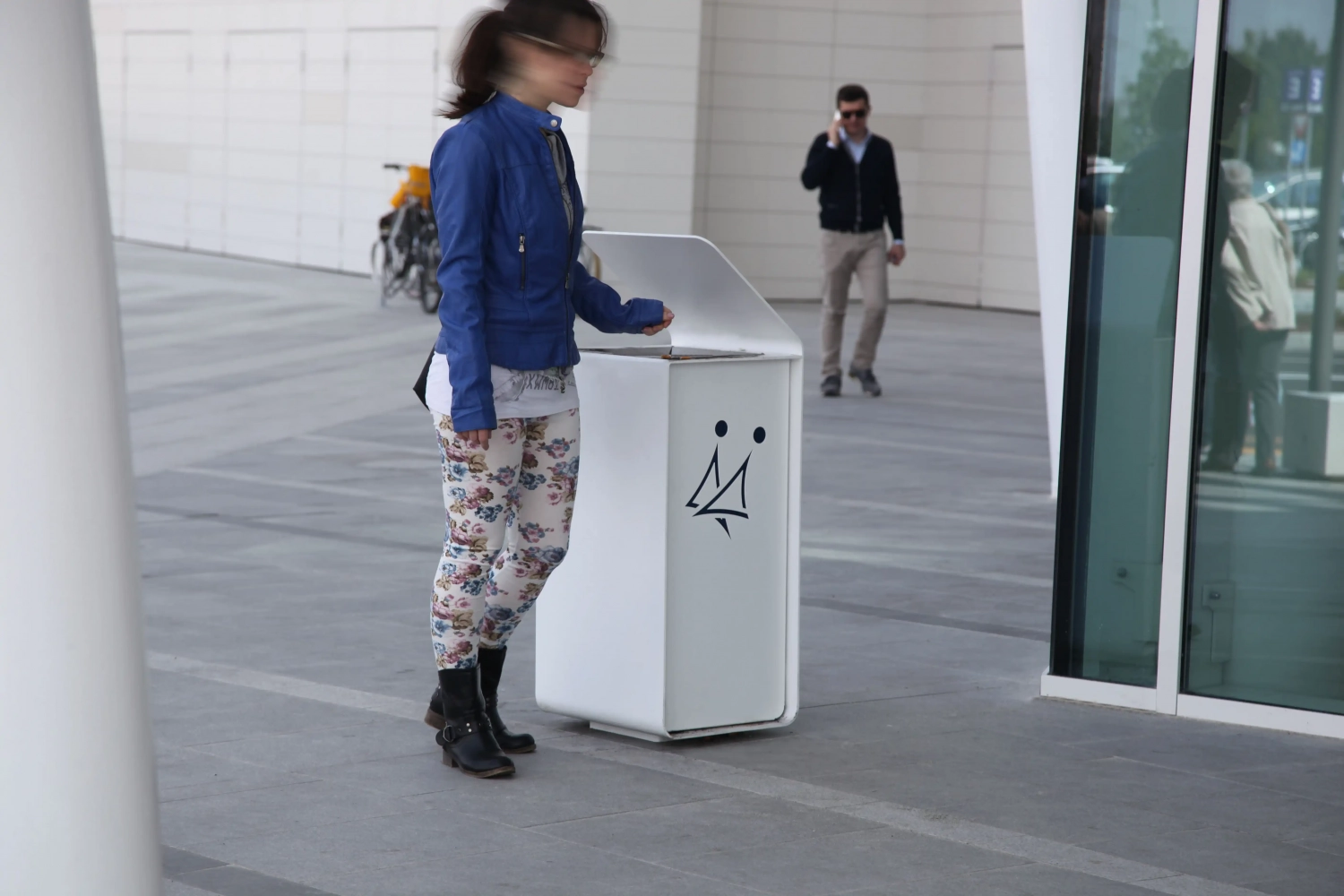 YES_1665 Eine Person in blauer Jacke und gemusterten Leggings steht neben einem weißen Abfallbehälter mit einem Gesichtsmotiv. Im Hintergrund ist eine weitere Person zu sehen, die telefoniert.