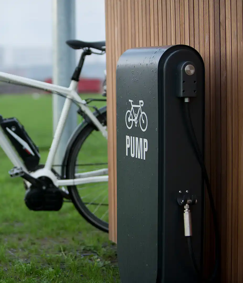 Alt-Text: "Fahrradpumpe mit Schlauch, auf einer Wiese neben einem Fahrrad, vor einer Holzverkleidung."