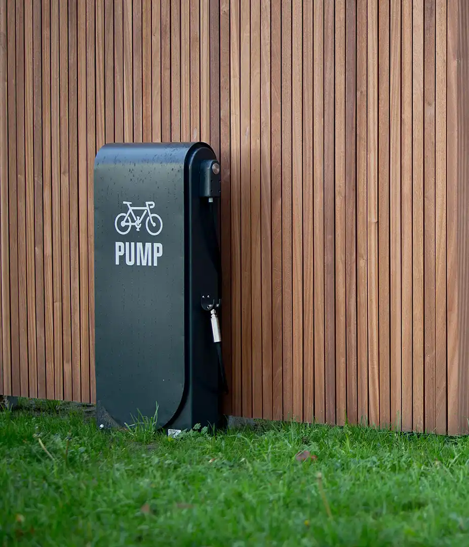 Alt-Text: Schwarze Fahrradpumpe mit Fahrrad-Symbol, aufgestellt neben einer Holzverkleidung in einem grünen Umfeld.