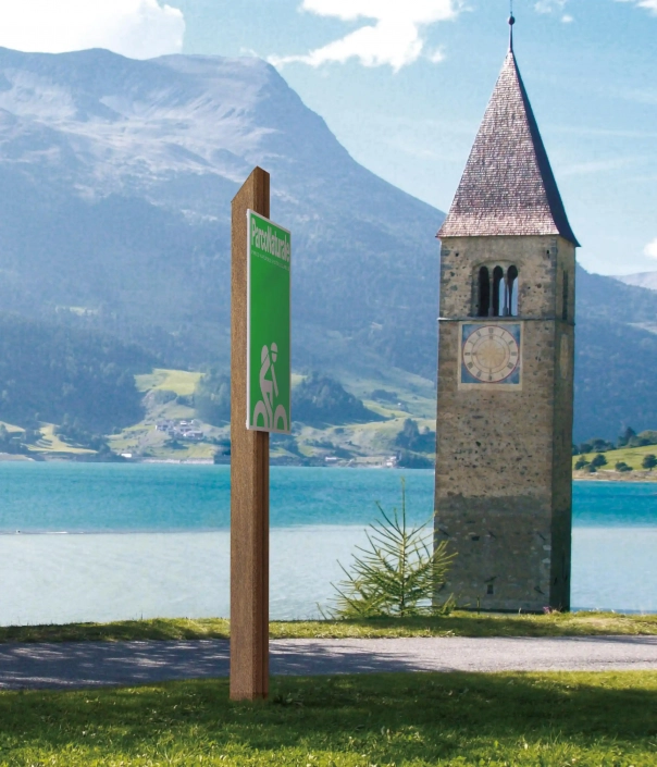 aspen-hinweistafel-02 Alt-Text: Hinweisschild für Radfahrer in der Nähe eines Sees, mit einem historischen Kirchturm im Hintergrund und Bergen in der Ferne.
