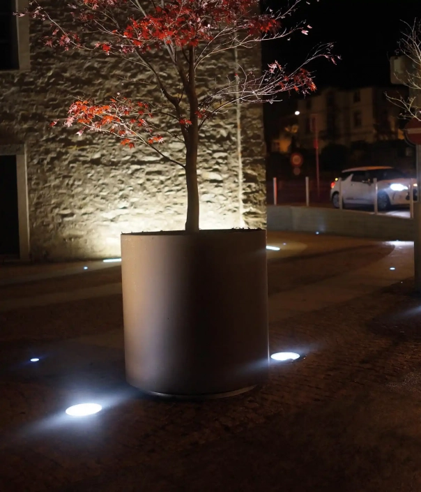 Alt-Text: Pflanzbehälter in der Nacht, beleuchtet von umgebenden Bodenlichtern, vor einer natursteinernen Wand.