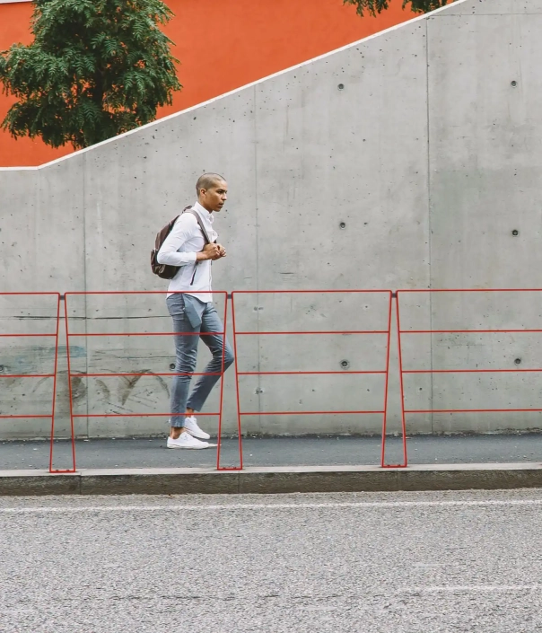 Alt-Text: Eine Person geht entlang einer roten Absperrung auf einem Gehweg, neben einer grauen Wand und einem orangefarbenen Hintergrund.