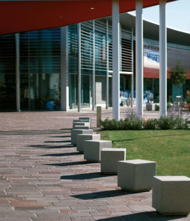 Eine Reihe von quadratischen Pollern auf einem gepflasterten Weg vor einem modernen Gebäude, umgeben von Grünflächen.