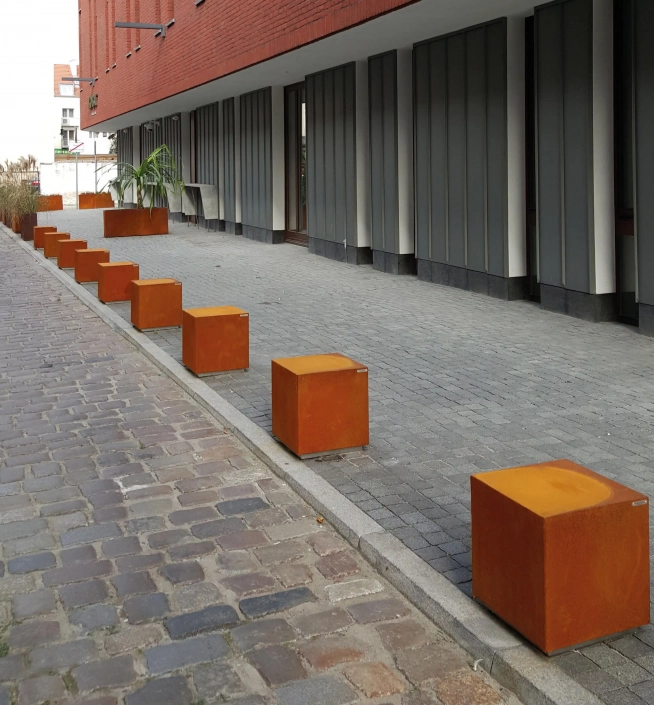 Eine Reihe von quadratischen, rostfarbenen Pollern auf einem Gehweg vor einem modernen Gebäude, umgeben von grauen Pflastersteinen.