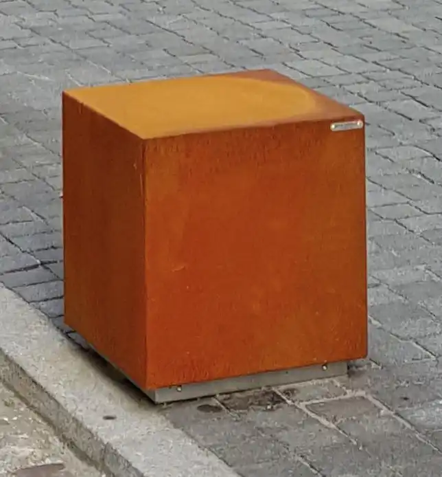 Alt-Text: Ein quadratischer, rostfarbener Poller aus Metall steht auf einem gepflasterten Gehweg.