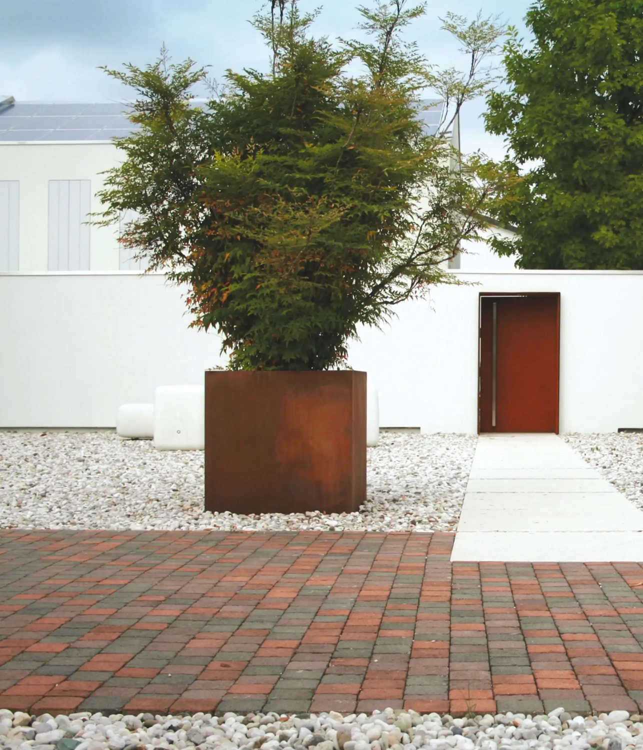 Alt-Text: Ein quadratischer, rustikaler Pflanzbehälter aus Metall, der einen großen Baum enthält, steht in einem modernen Garten mit Kies und roten Pflastersteinen. Im Hintergrund ist eine weiße Wand und eine Tür sichtbar.