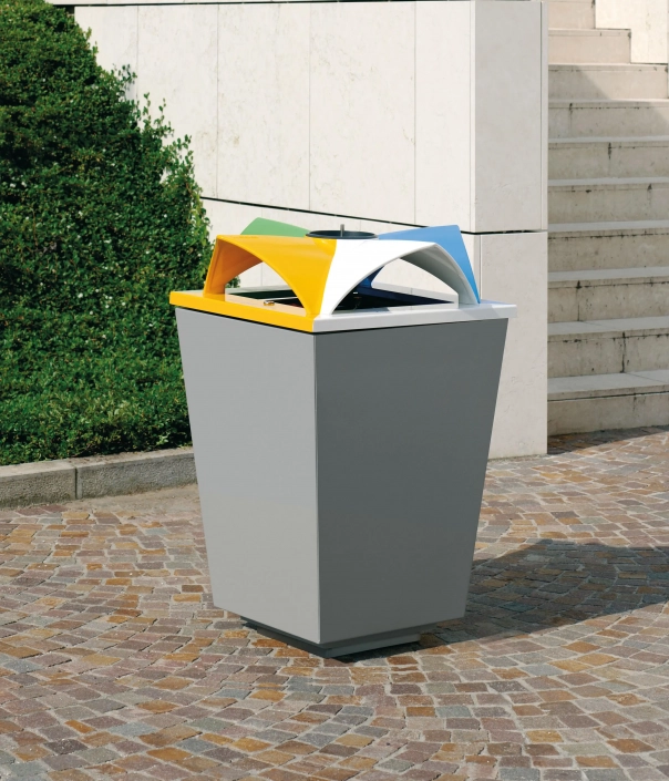 Ein moderner Abfallbehälter mit farbigen Deckeln für die Mülltrennung steht auf einem gepflasterten Platz. Im Hintergrund sind eine grüne Hecke und Treppenstufen sichtbar.