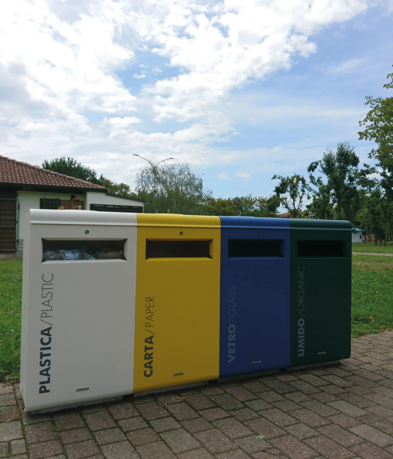 Abfalltrennung mit vier Recycling-Containern in einer grünen Parklandschaft: blau für Plastik, gelb für Papier, grün für organische Abfälle und grau für Glas. Im Hintergrund ist ein Gebäude und Bäume sichtbar.