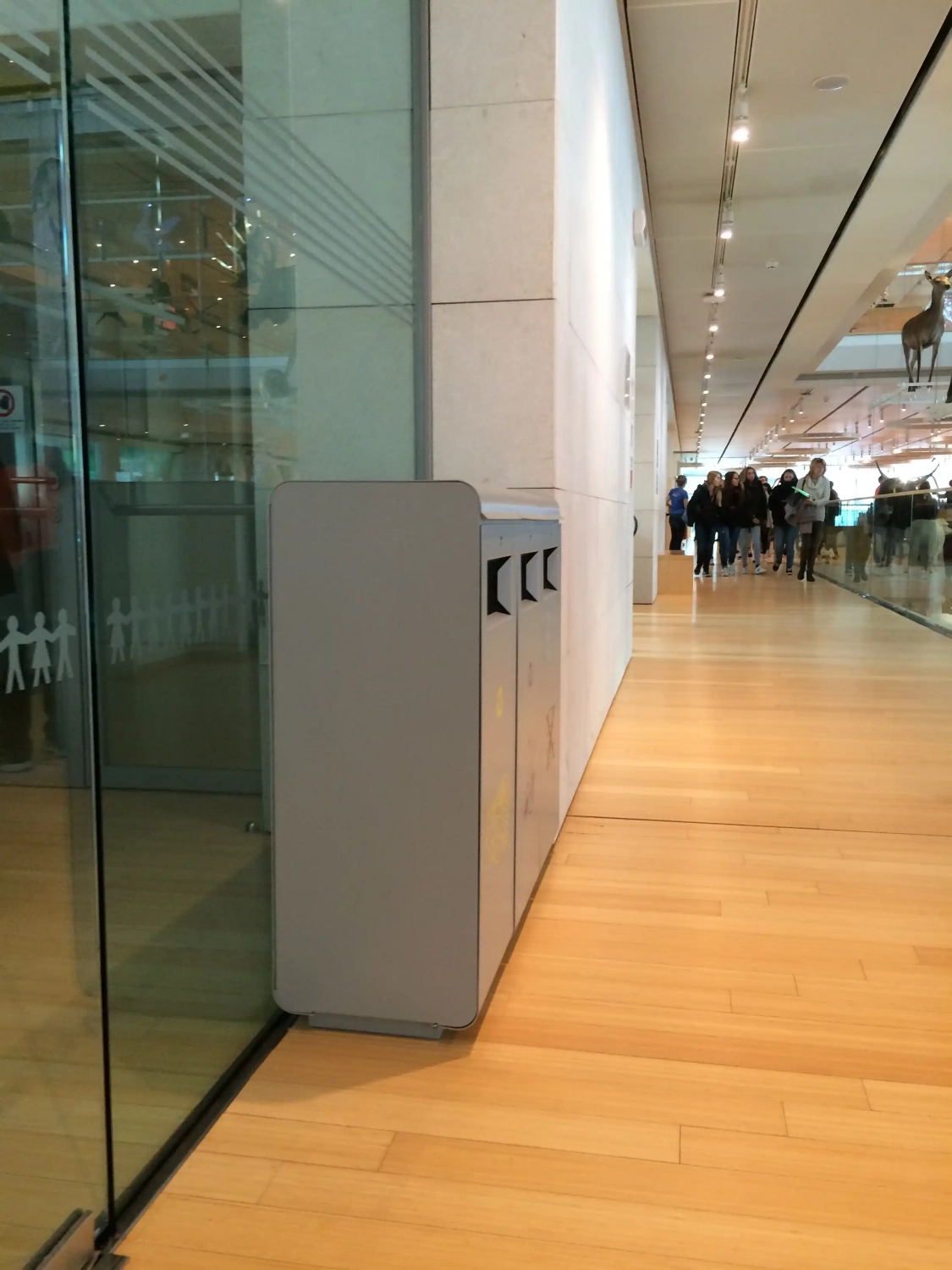 Ein grauer Abfalleimer steht an einer Wand aus Glas in einem modernen Innenraum, während Menschen im Hintergrund entlang eines Holzfußbodens gehen.