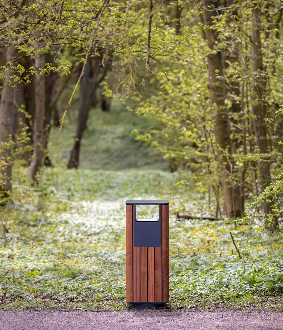 Ein moderner Abfalleimer aus Holz steht in einem grünen Waldgebiet, umgeben von Bäumen und frischem Laub. Der Weg hinter dem Eimer führt tiefer in den Wald.