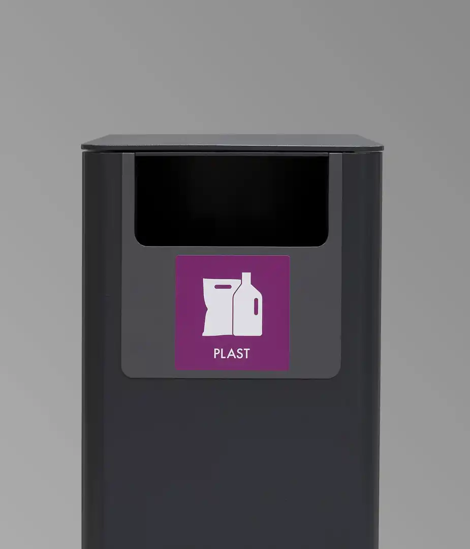 Ein schwarzer Müllcontainer mit einer beschrifteten Öffnung für Plastikmüll, gekennzeichnet durch ein lila Schild mit einem Symbol für Plastikflaschen und -tüten.