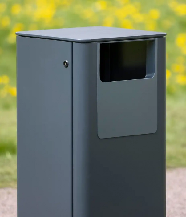 Ein grauer, moderner Abfallbehälter steht auf einem Weg, umgeben von gelben Blumen.