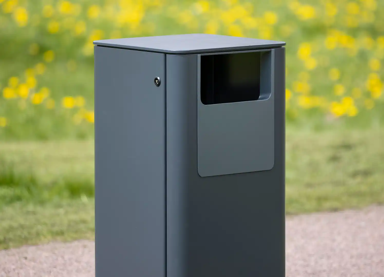 Eine moderne, graue Abfalltonne steht auf einem Weg, im Hintergrund leuchtet eine Wiese mit gelben Blumen.
