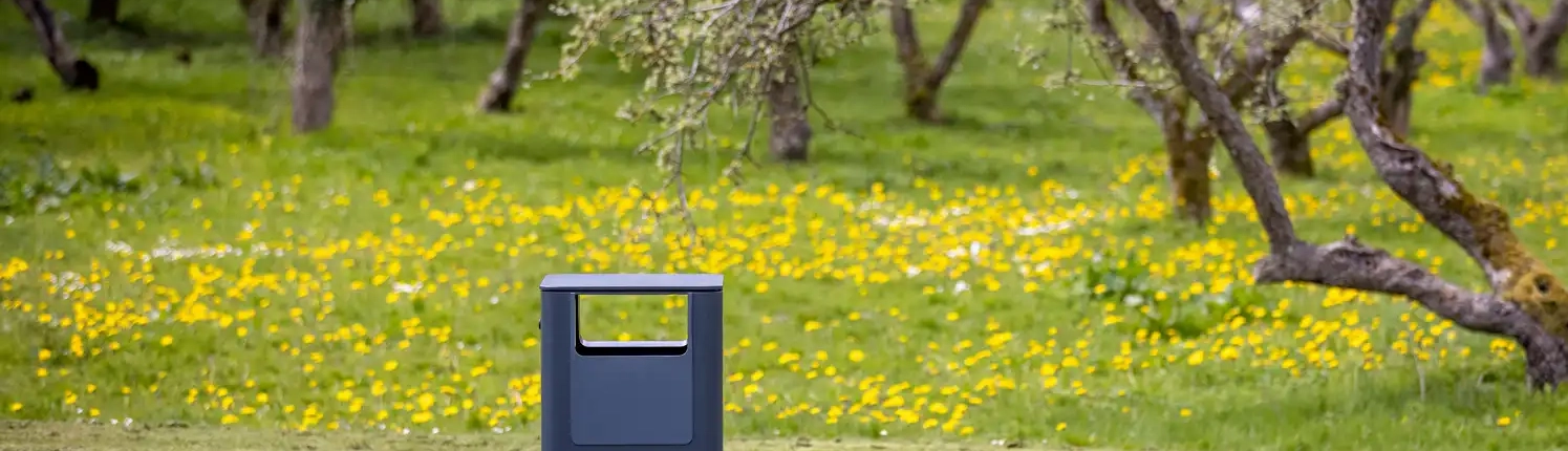 egedal-2-abfallbehaelter-07 Müllbehälter auf einem Weg in einer blühenden Parklandschaft mit Bäumen und gelben Blumen auf dem Gras.