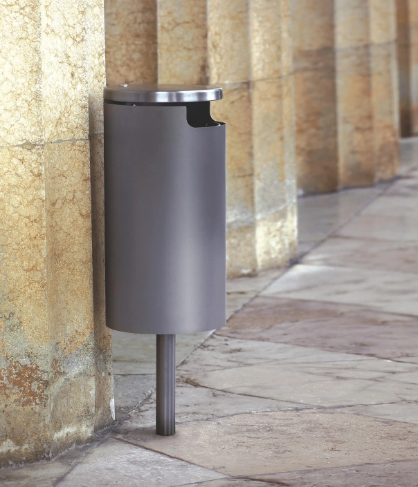 Ein moderner, runder Abfallbehälter aus Metall, der an einer Steinmauer steht. Der Behälter hat einen offenen oberen Rand und ist auf einem stabilen Standfuß montiert.