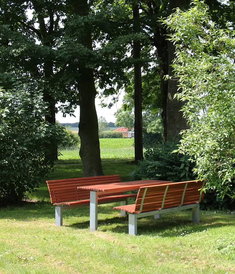 Alt-Text: Eine rustikale Sitzgruppe aus Holz und Metall steht in einem begrünten urbanen Raum, umgeben von Bäumen und Sträuchern, ideal für soziale Begegnungen und Entspannung im Freien.