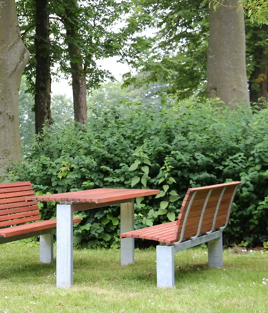 Alt-Text: Moderne Sitzgruppe im urbanen Freiraum, bestehend aus einem Tisch und zwei Bänken, umgeben von dichtem Grün.