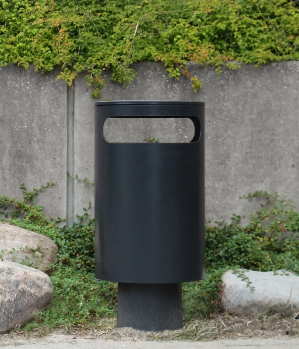 Ein schwarzer Müllcontainer steht vor einer grauen Wand, umgeben von Grünpflanzen und Steinen.