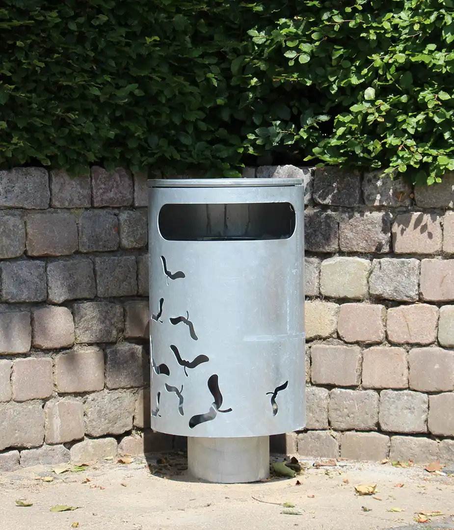 Ein moderner, silberner Abfallbehälter mit Ausschnitten, der an einer Steinmauer steht, umgeben von grünen Sträuchern.