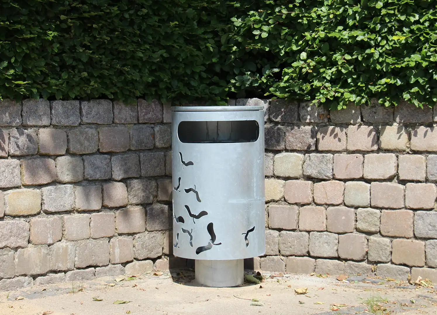 Ein silberner Müllcontainer mit einem schlichten Design, das von einer mit Blättern bewachsenen Mauer umgeben ist. Auf der Oberfläche des Containers sind stilisierte Vogelmotive angebracht.