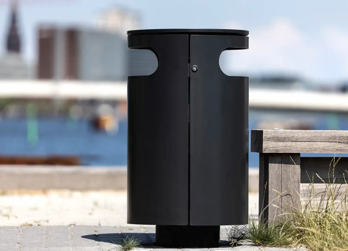 Alt-Text: Ein moderner, schwarzer Müllcontainer steht am Ufer eines Gewässers, im Hintergrund sind eine Brücke und städtische Gebäude zu erkennen.