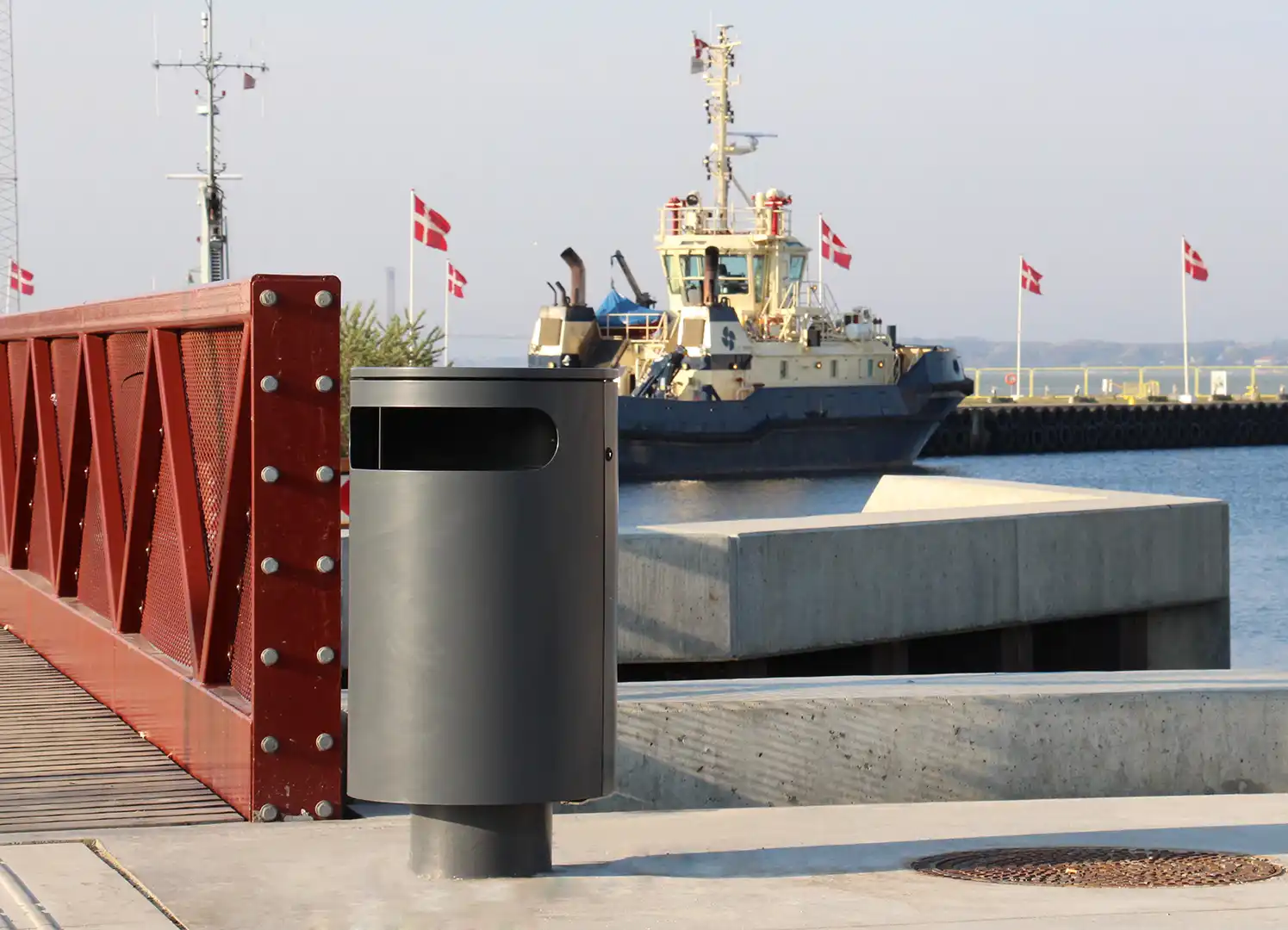 Ein moderner Mülleimer steht auf einer Hafenpromenade, im Hintergrund ist ein Schiff mit dänischer Flagge zu sehen.