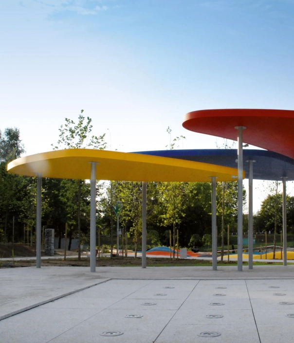 Alt-Text: Bunte Überdachungen in einer Freifläche, unterstützt von silbernen Säulen, umgeben von Bäumen und einem Spielplatz.