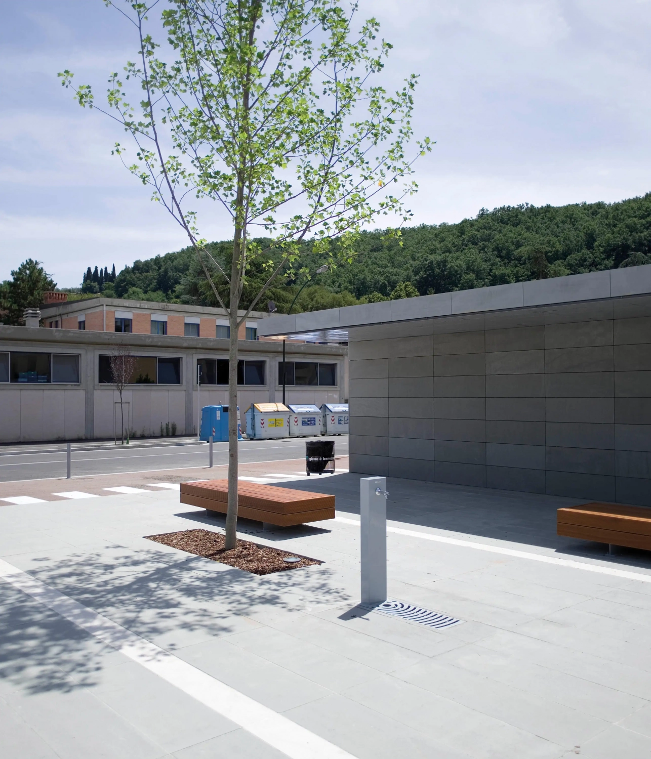 Alt-Text: Urbaner Raum mit Sitzgruppen aus Holz, einem Trinkwasserbrunnen und einem kleinen Baum, sowie einer modernen Gebäude-Fassade im Hintergrund.