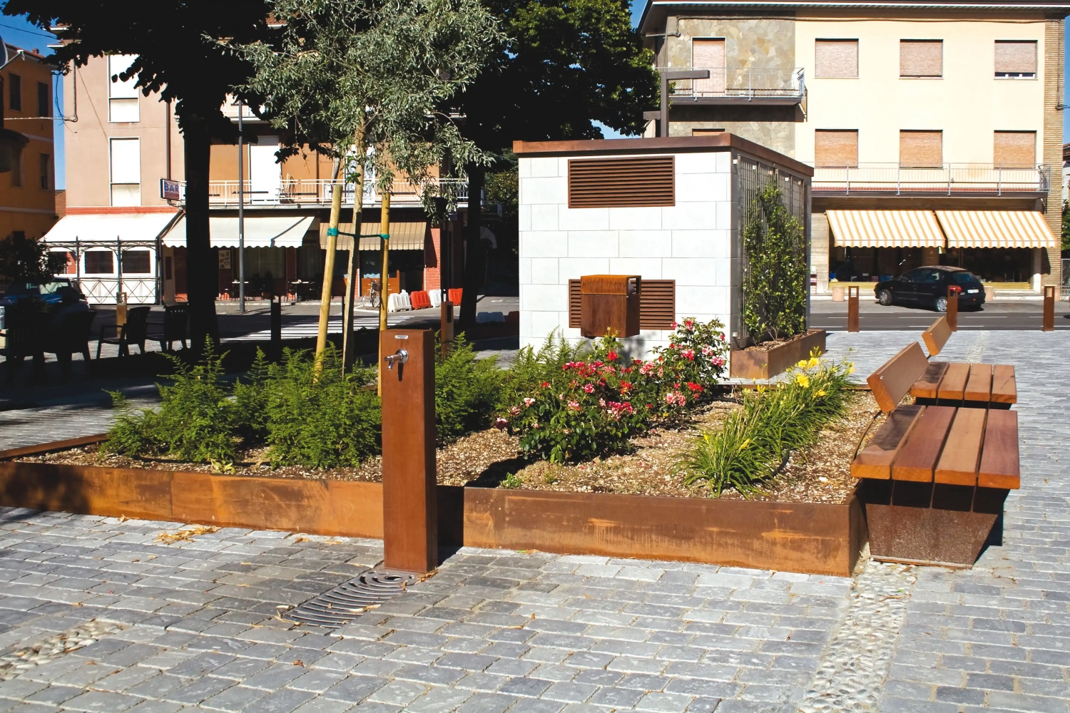 Alt-Text: Urbaner Raum mit Sitzgruppen aus Holz, einem Trinkwasserbrunnen und bepflanzten Beeten, umgeben von Gebäuden.