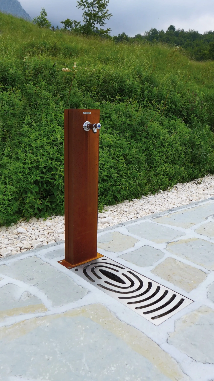 Alt-Text: "Ein moderner Trinkwasserbrunnen aus rostfreiem Stahl, umgeben von einer gepflegten Grünfläche und einem gepflasterten Boden."