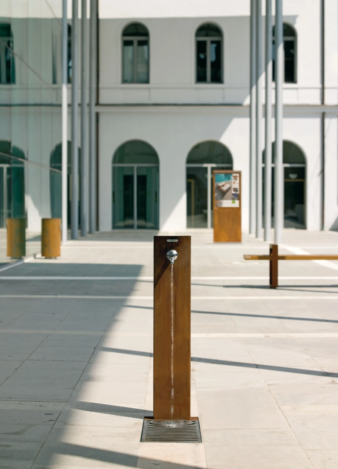 Alt-Text: Urbaner Raum mit modernen Sitzgruppen, einem Wasserspender und Grillbereichen, umgeben von einer architektonisch ansprechenden Kulisse mit Glasfronten.