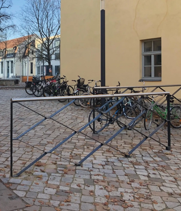 Alt-Text: "Metallene Absperrungen auf einem gepflasterten Platz mit mehreren abgestellten Fahrrädern im Hintergrund."