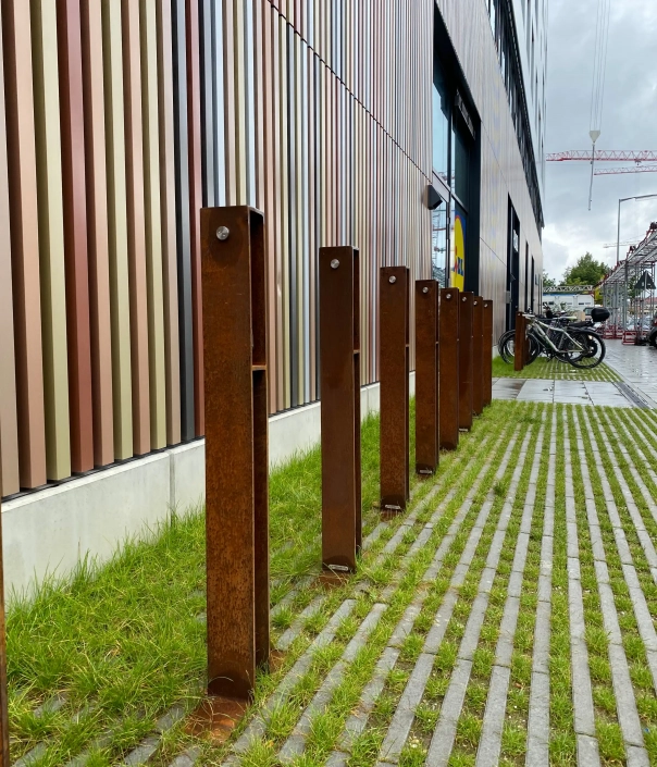 guardia-radverkehr-09 Alt-Text: "Reihe aus rostigem Fahrradständern neben einer modernen Gebäudefassade, umgeben von Rasengräsern und einem gepflasterten Weg."