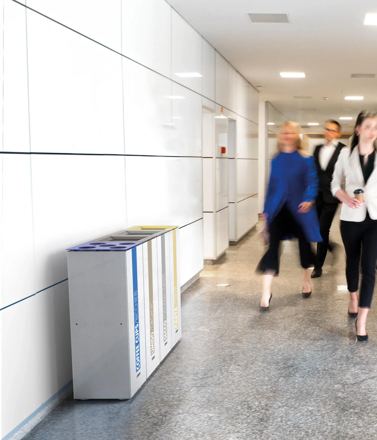 Ein geschäftiger Flur mit mehreren Personen, die in formeller Kleidung vorbeigehen. Einige tragen Kaffeebecher, während im Hintergrund Mülleimer sichtbar sind.