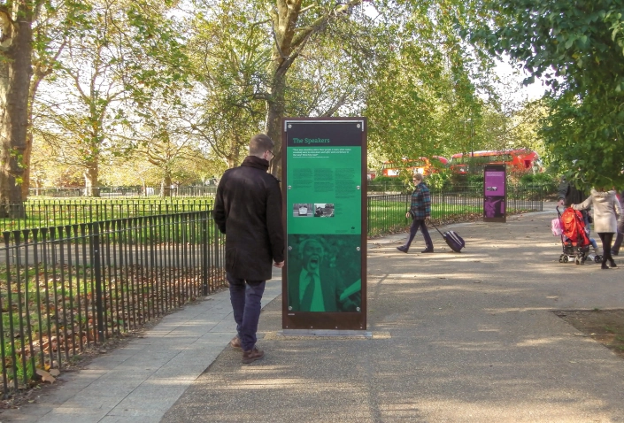 icaro-hinweistafel-09 Alt-Text: Ein Besucher steht vor einem Hinweisschild im Park, während andere Menschen im Hintergrund spazieren gehen. Das Schild informiert über die Geschichte und Bedeutung eines besonderen Ortes.