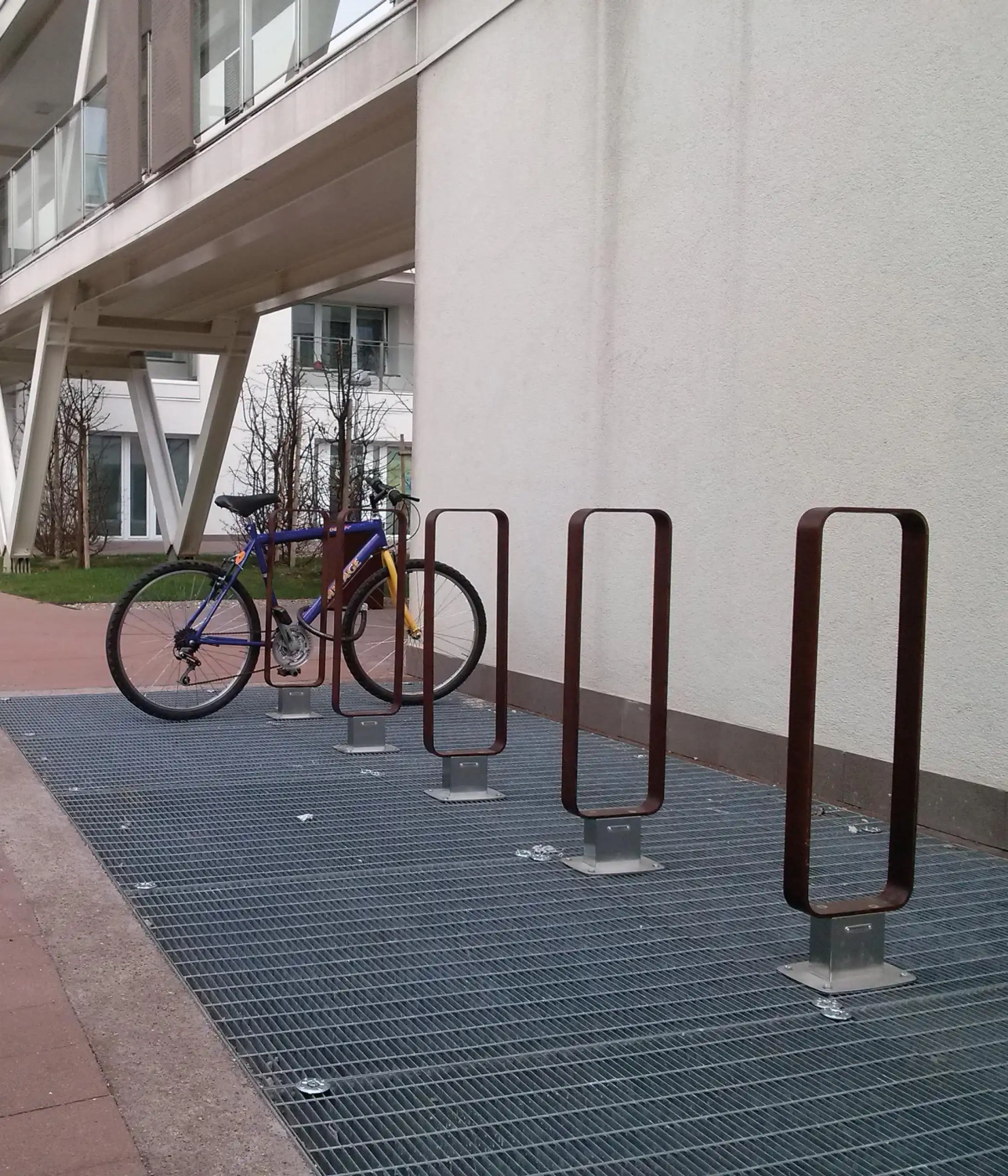 Alt-Text: "Fahrradständer aus Edelstahl mit einem abgestellten Fahrrad auf einem gepflasterten Bereich vor einem modernen Gebäude."