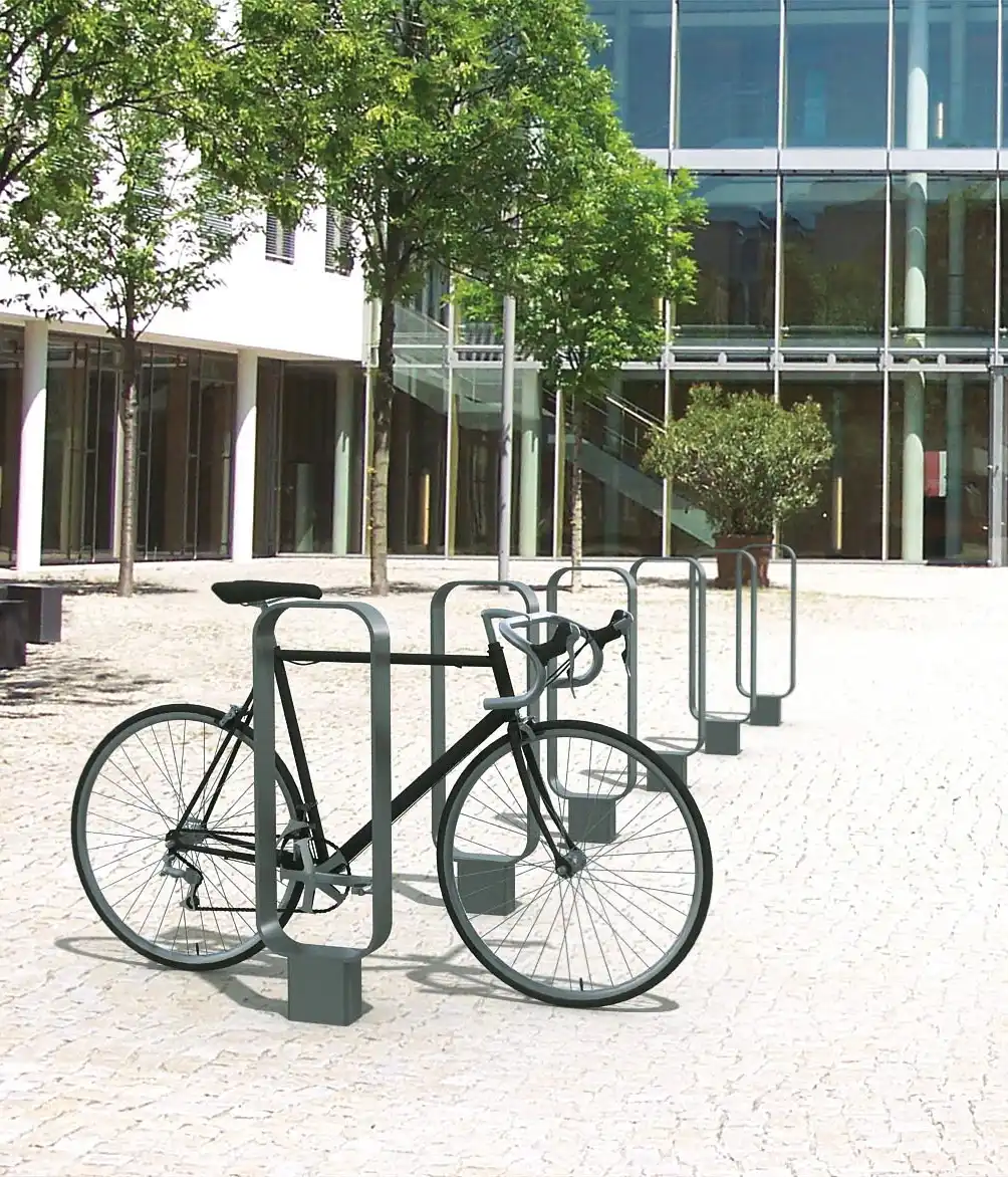 Alt-Text: "Moderne Fahrradständer in einer urbanen Umgebung mit einem Fahrrad, umgeben von Bäumen und modernen Gebäuden."
