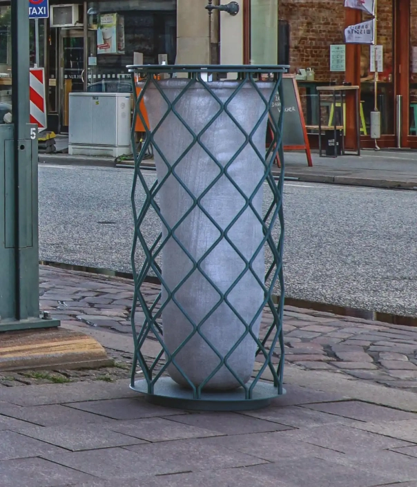 Ein zylindrischer Müllbehälter steht in einem geschützten Gitterbehälter auf einem Gehweg, umgeben von einer städtischen Umgebung mit Geschäften und einem Bürgersteig.