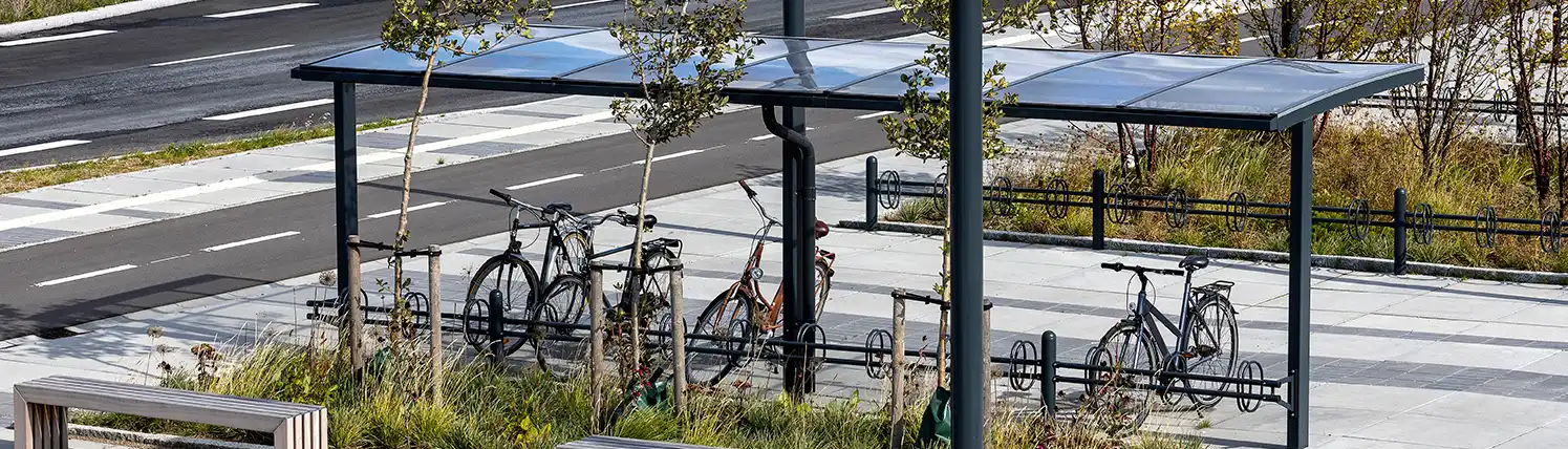 Alt-Text: "Moderne Überdachung für Fahrräder mit mehreren abgestellten Fahrrädern darunter, umgeben von einer grünen Umgebung und einem modernen Straßenbelag."